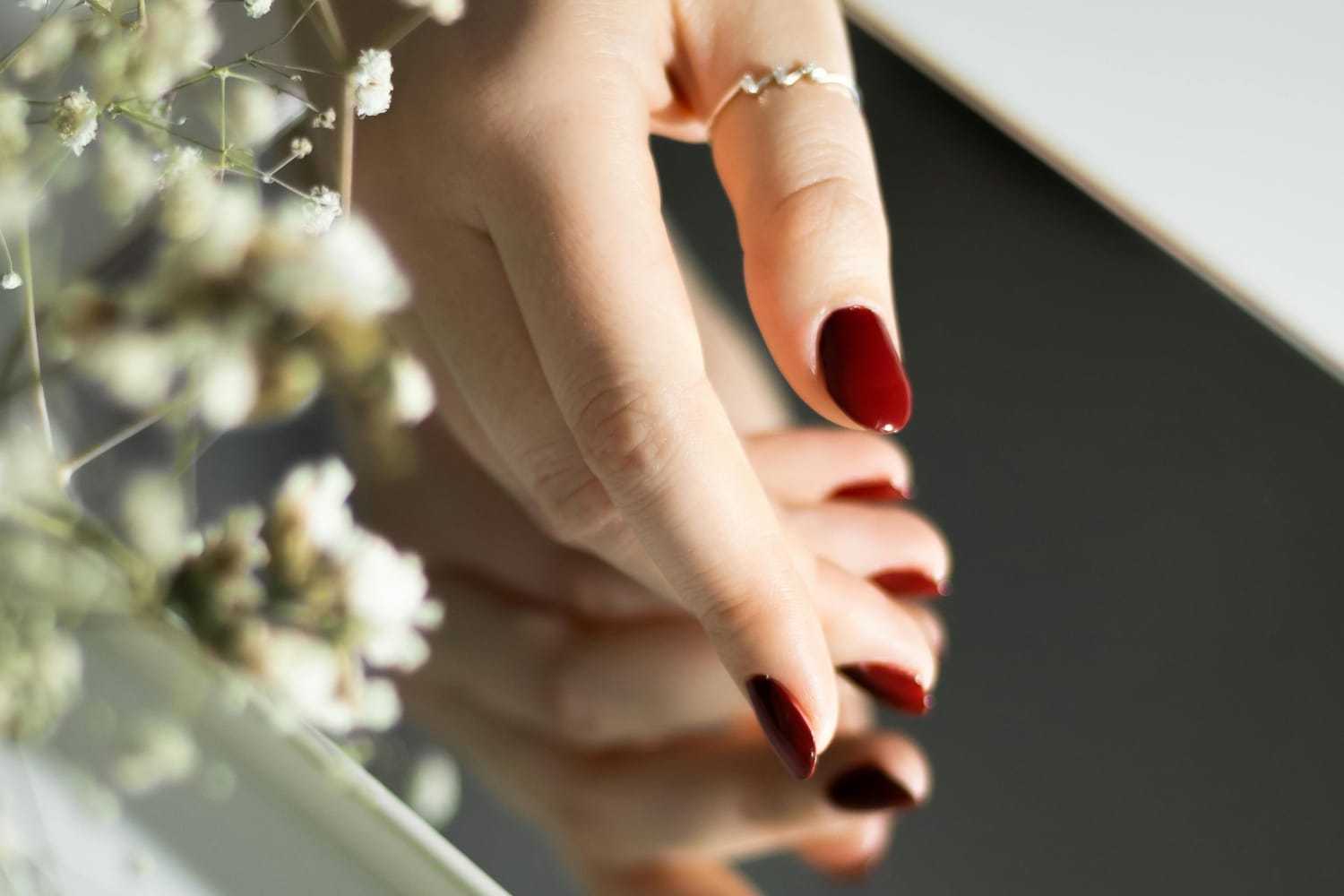 Hand with red nails and a ring reflected in a mirror near delicate white flowers.